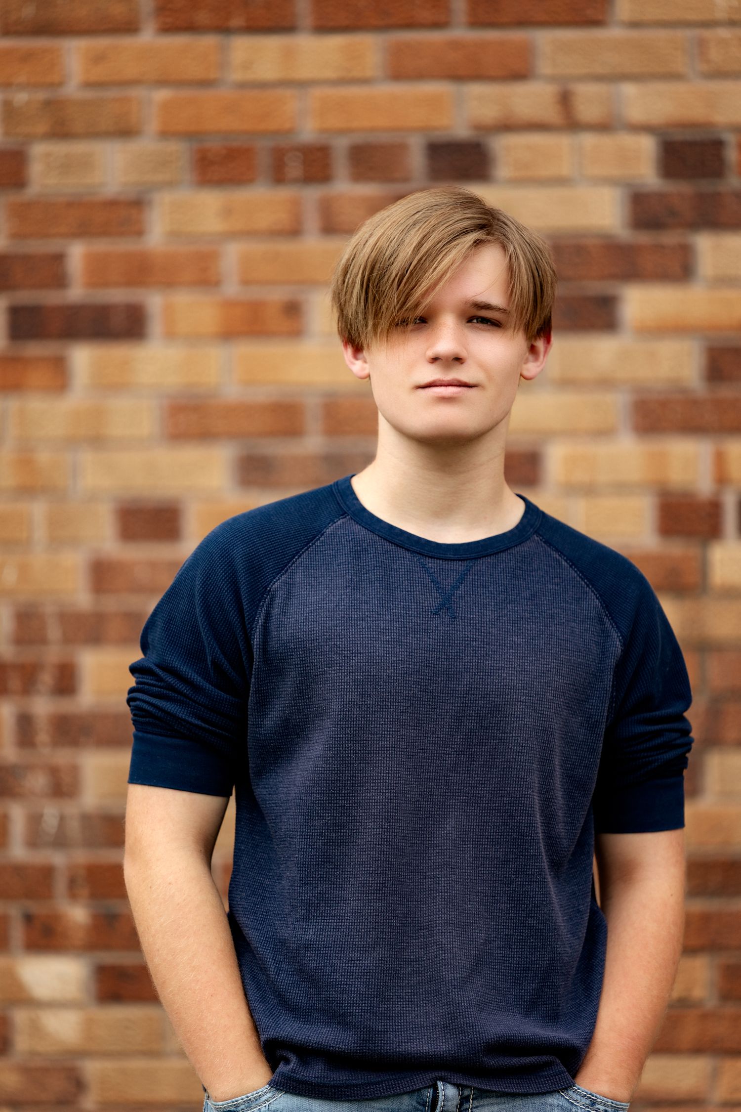 A teenage boy in a navy blue raglan shirt stands confidently in front of a brick wall with hands in pockets.
