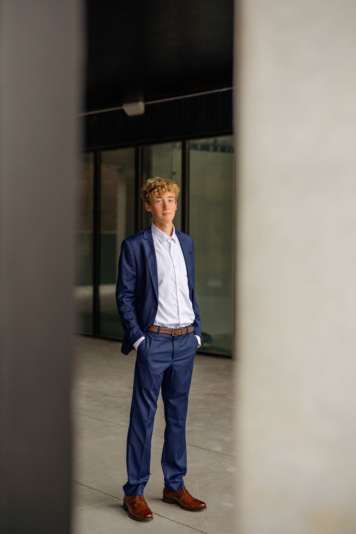 Young man in navy blue suit with white shirt and brown shoes standing confidently in modern building entrance.