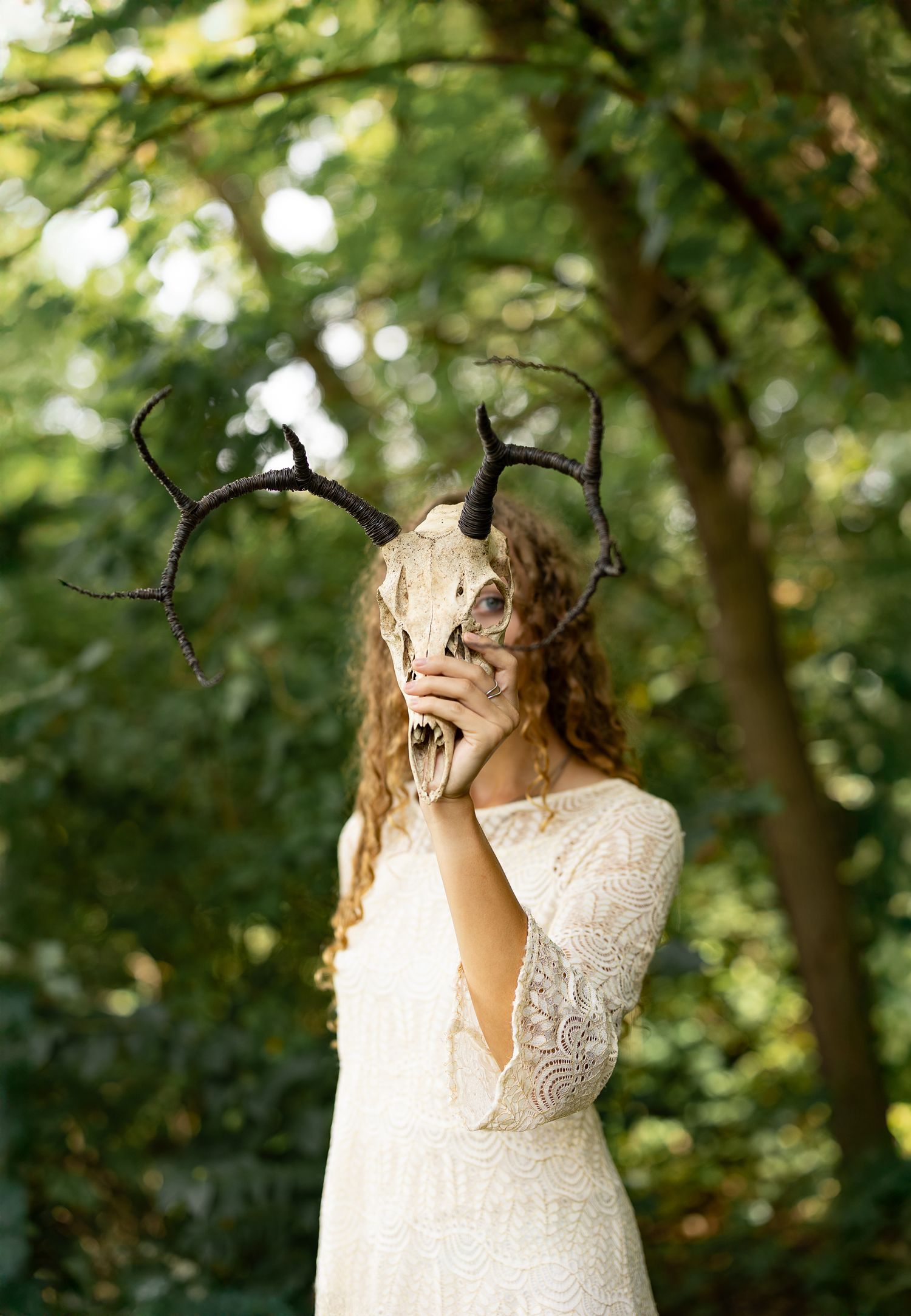 Woman in white lace dress holding a deer skull mask with antlers in a lush green forest setting.