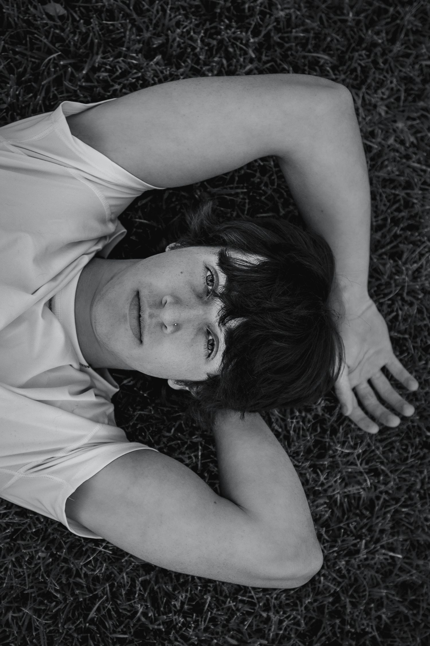 Black and white photo of a young person lying on grass, arms raised, looking up at the camera.