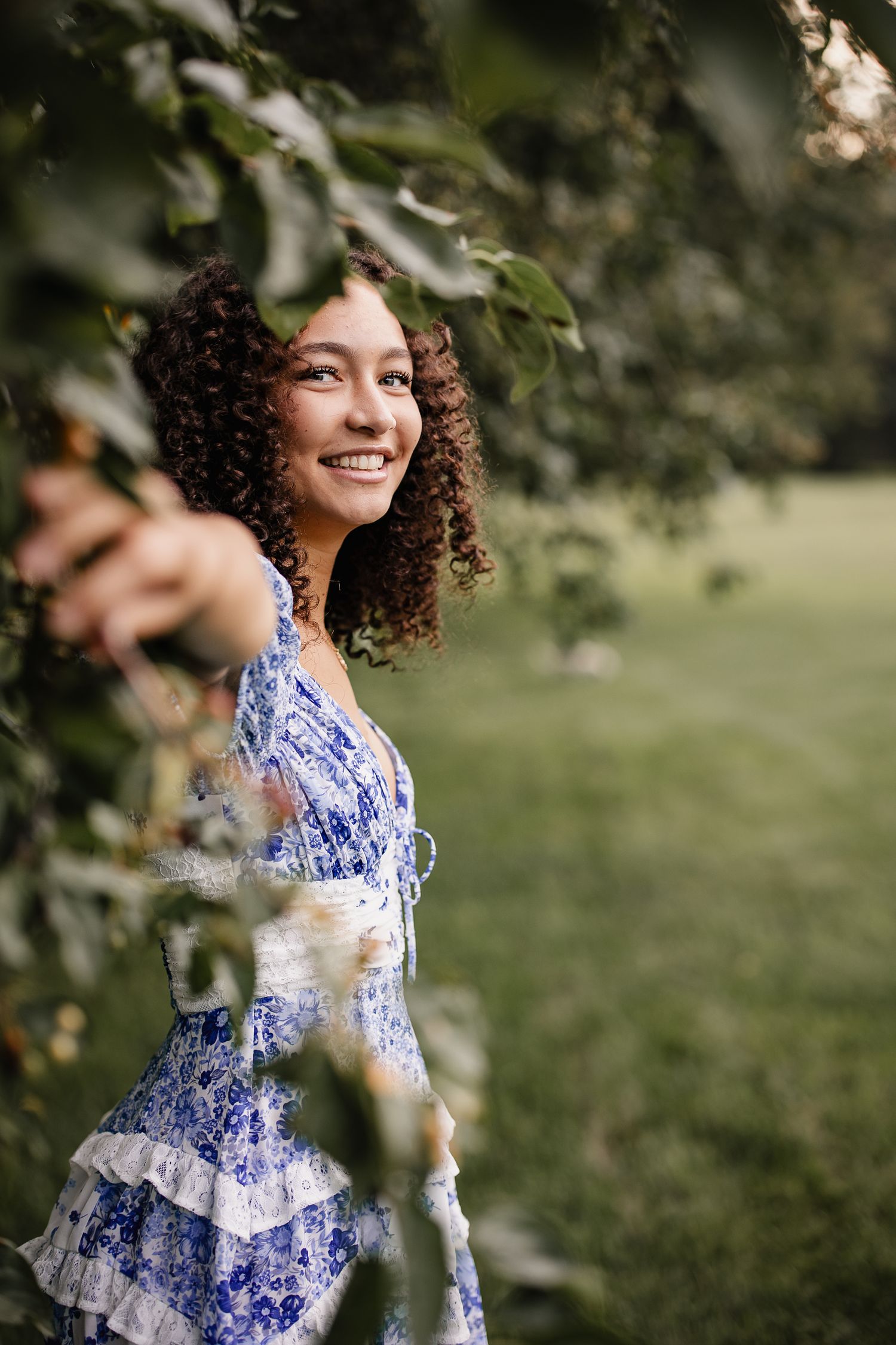 Smiling young woman with curly hair in a blue floral dress standing among green tree branches outdoors.