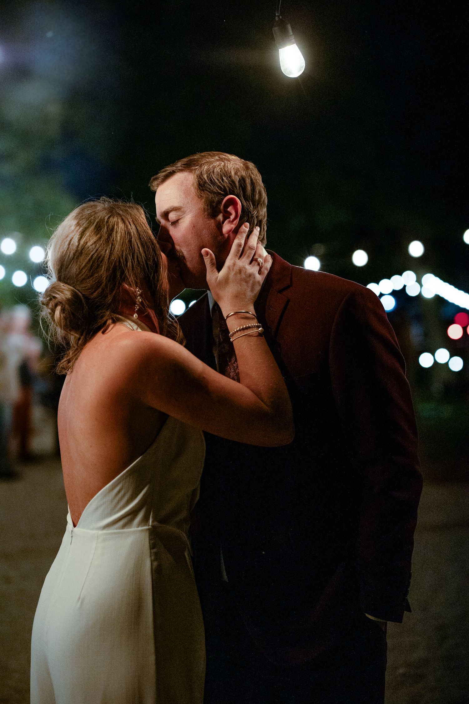 Bride and groom share a romantic kiss under string lights at an outdoor evening wedding reception.