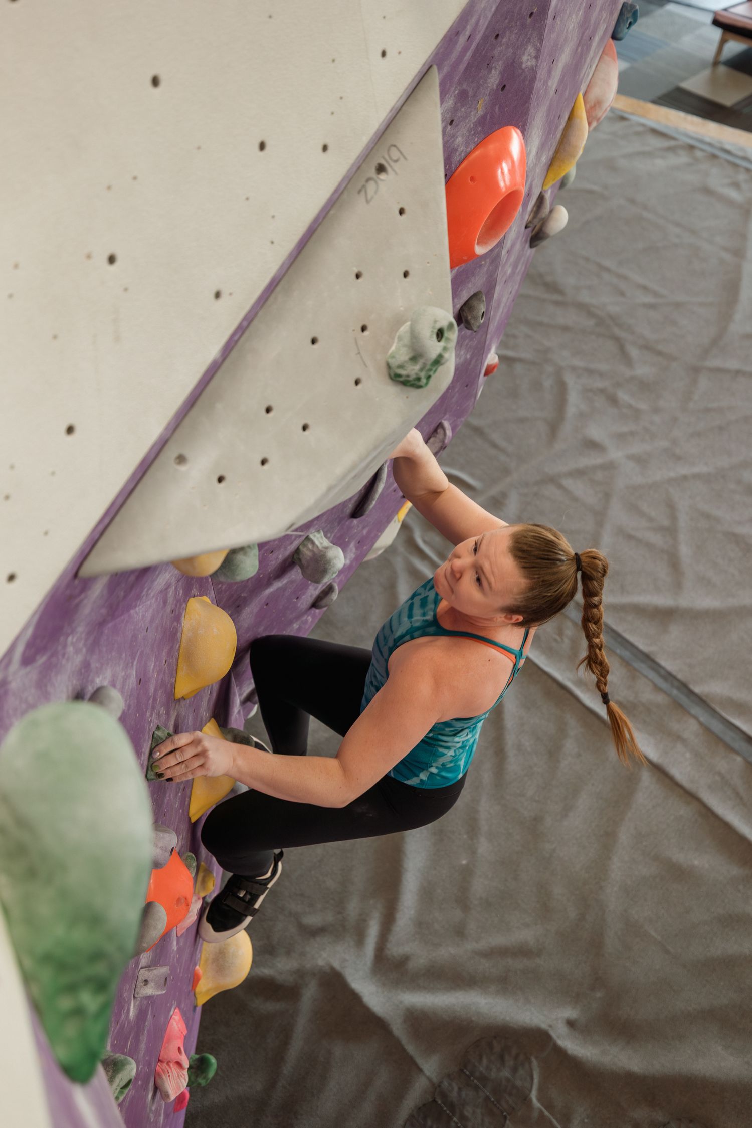 Woman rock climbing on colorful indoor bouldering wall, wearing teal top and black leggings, gripping holds.