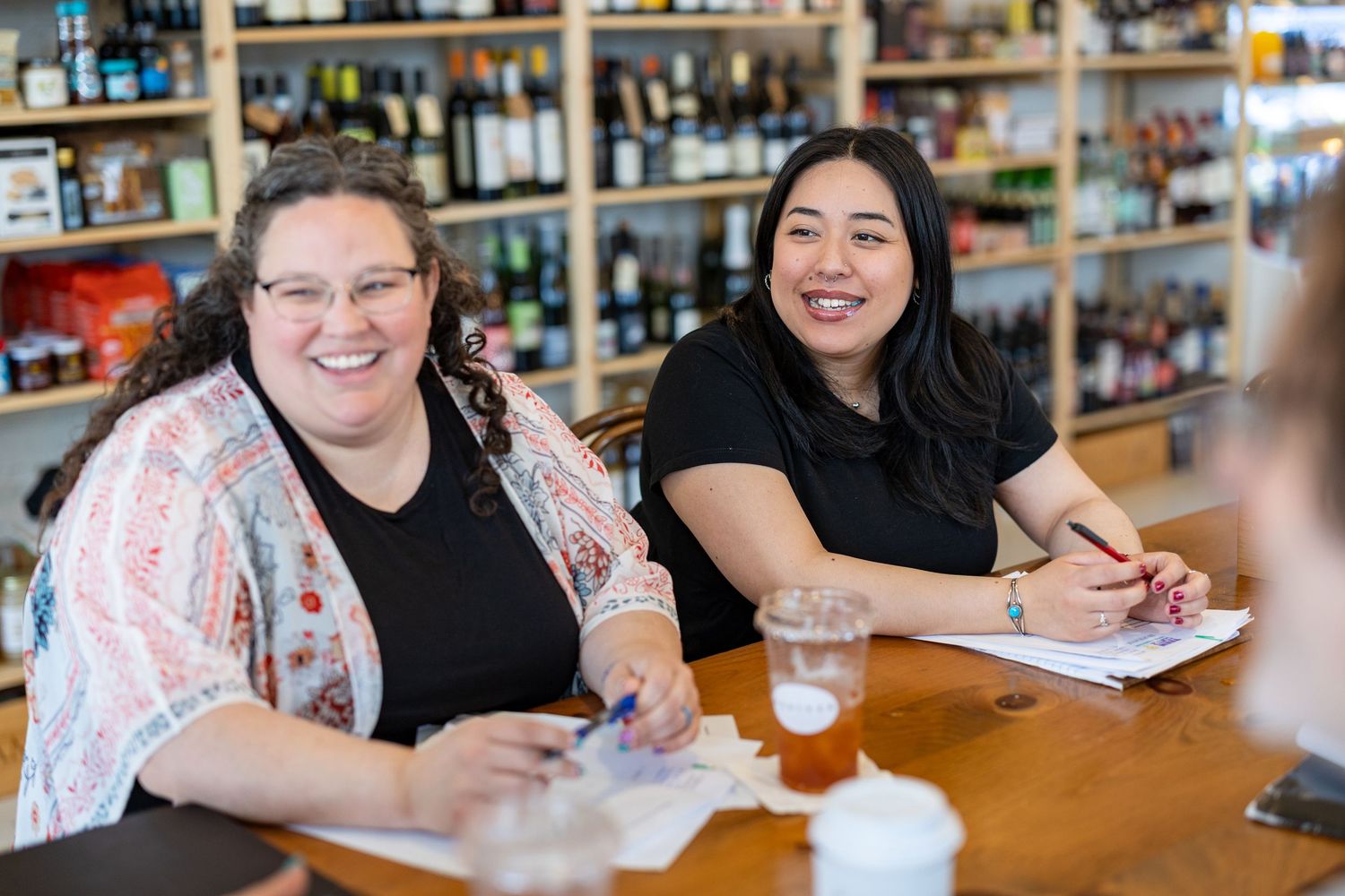 Two smiling women sitting at a bar counter with drinks, notebooks, and wine bottles displayed on shelves behind them.