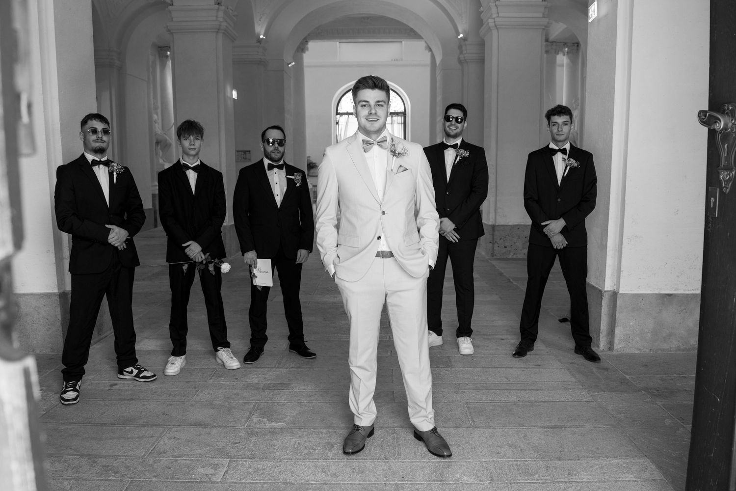 Groomsmen in tuxedos and white jackets posed formally in a classic interior.
