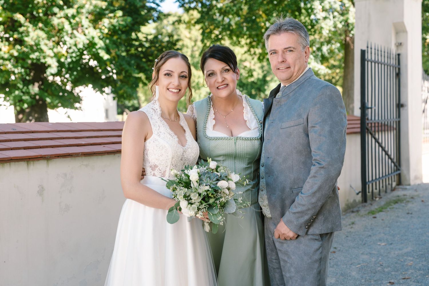 Wedding party poses outdoors under green trees near white pillars.