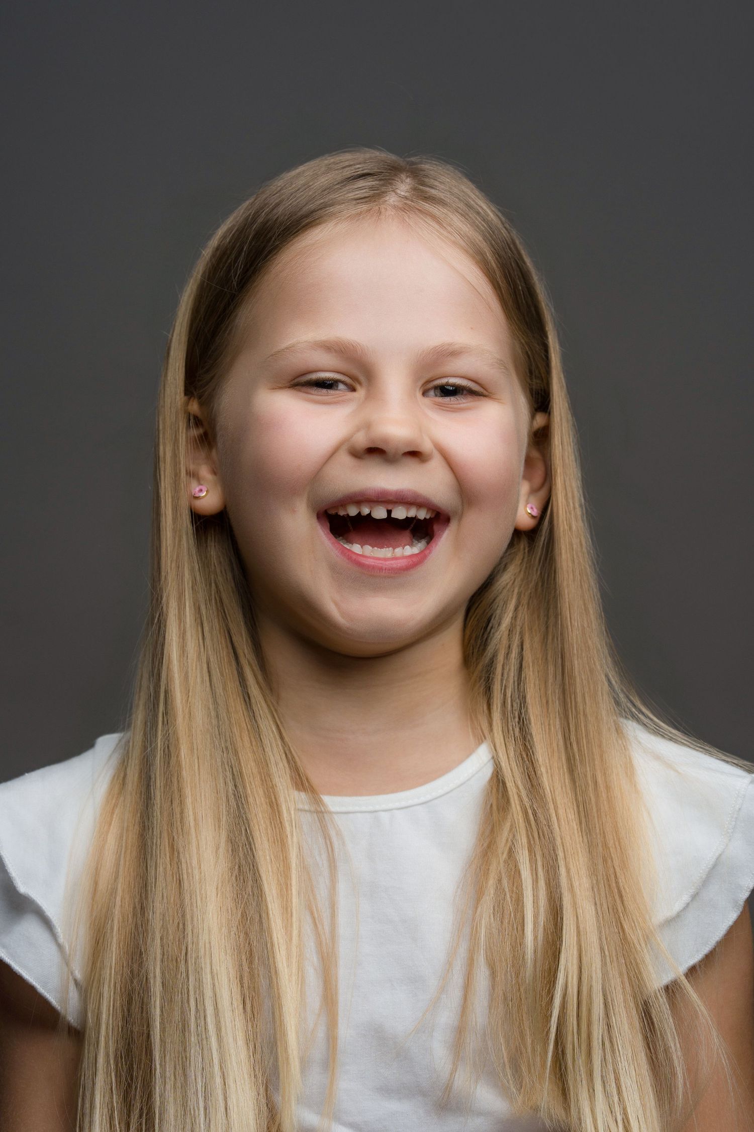 Portrait of a young child with long blonde hair wearing a white shirt and showing a big cheerful smile against a dark background.