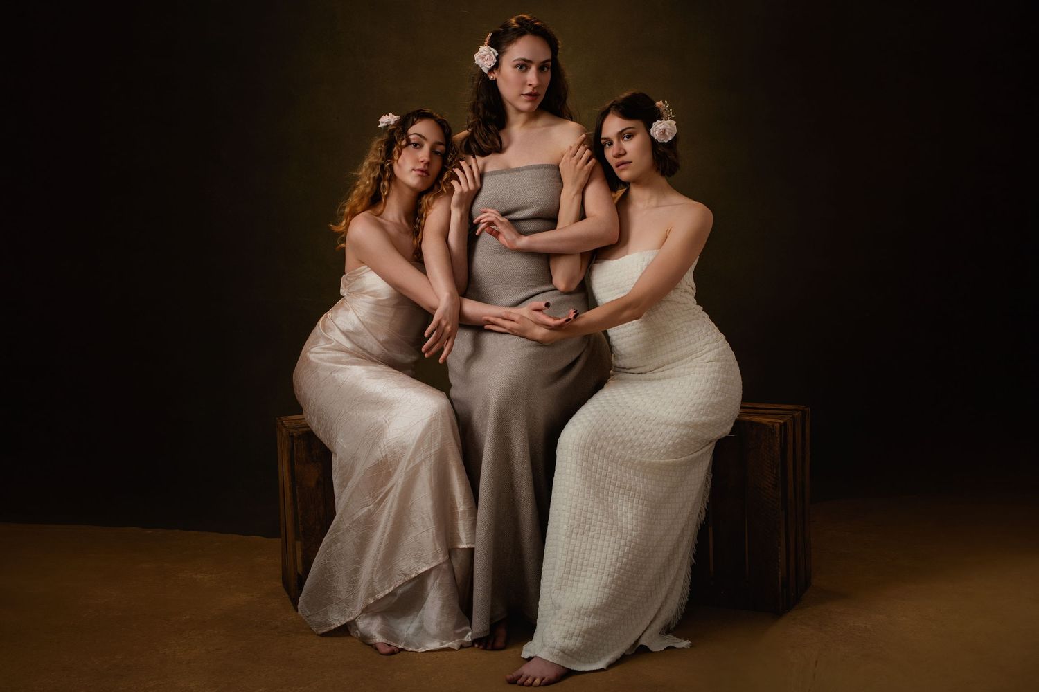 Three bridesmaids in flowing white dresses sit together on a vintage brown leather couch in a dark studio setting.