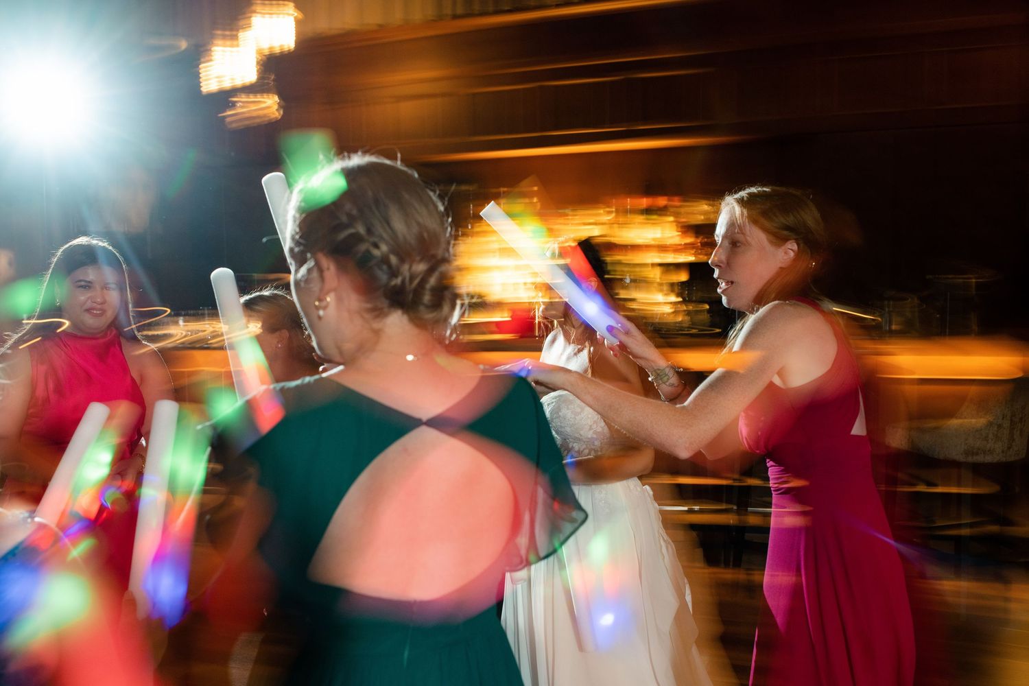Women in formal gowns dancing with colorful glow sticks at a lively wedding reception party.