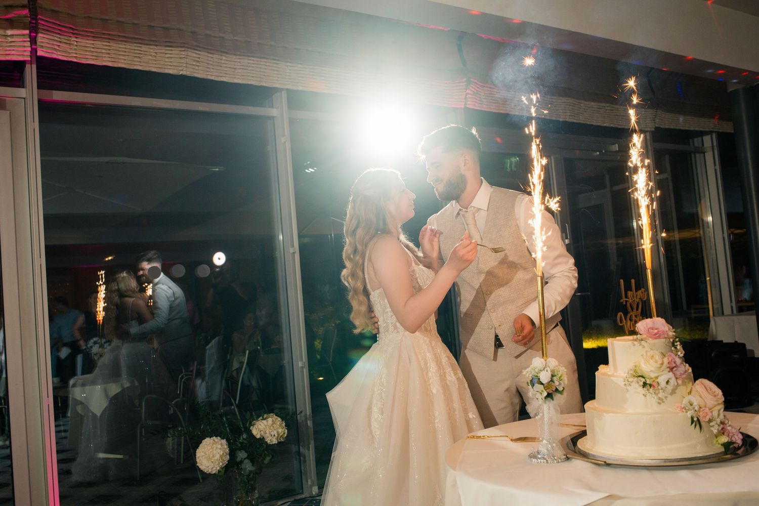 Bride and groom share a romantic kiss beside wedding cake with sparklers glowing at elegant evening reception.