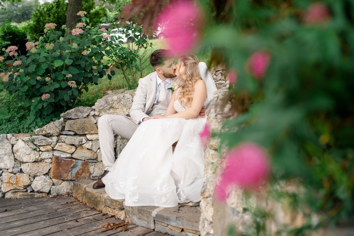 Bride and groom sharing a romantic moment seated on a stone wall surrounded by lush garden greenery and pink flowers.