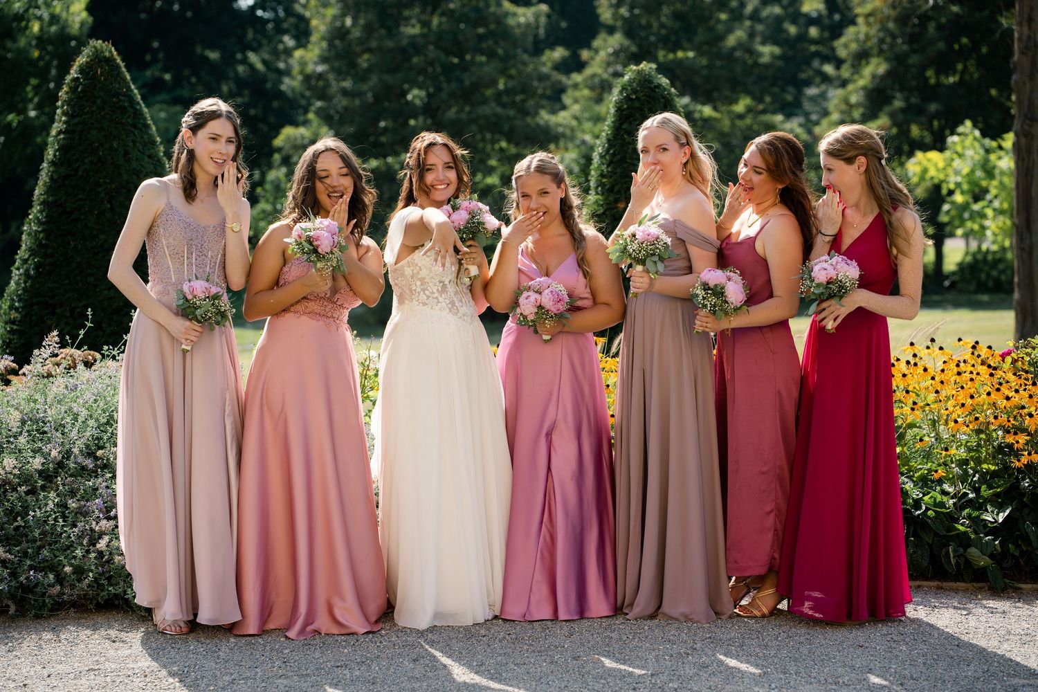 Bridesmaids in varying shades of pink dresses pose together in a garden.