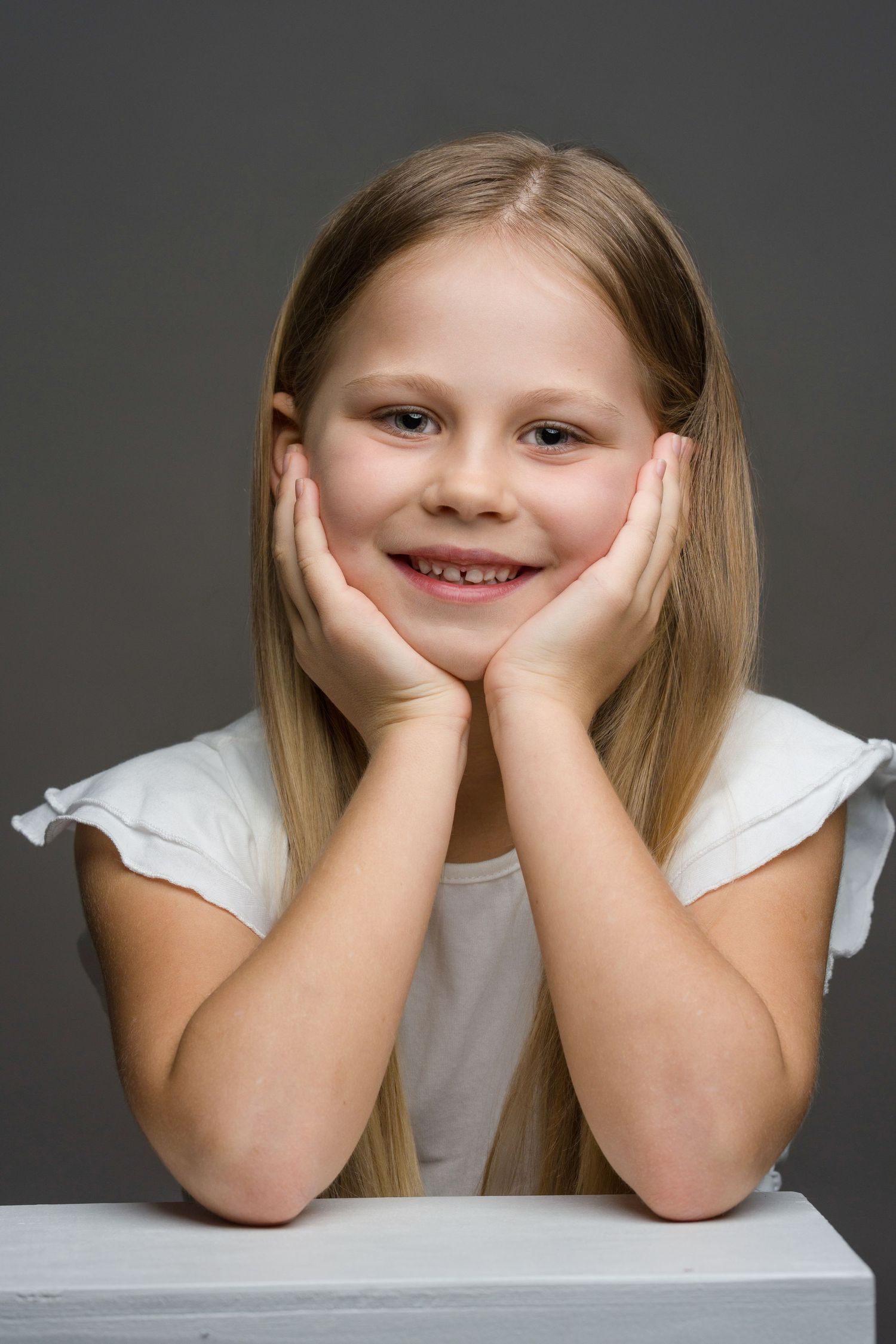 A young child with long blonde hair wearing a white shirt rests their head in their hands while smiling at the camera.