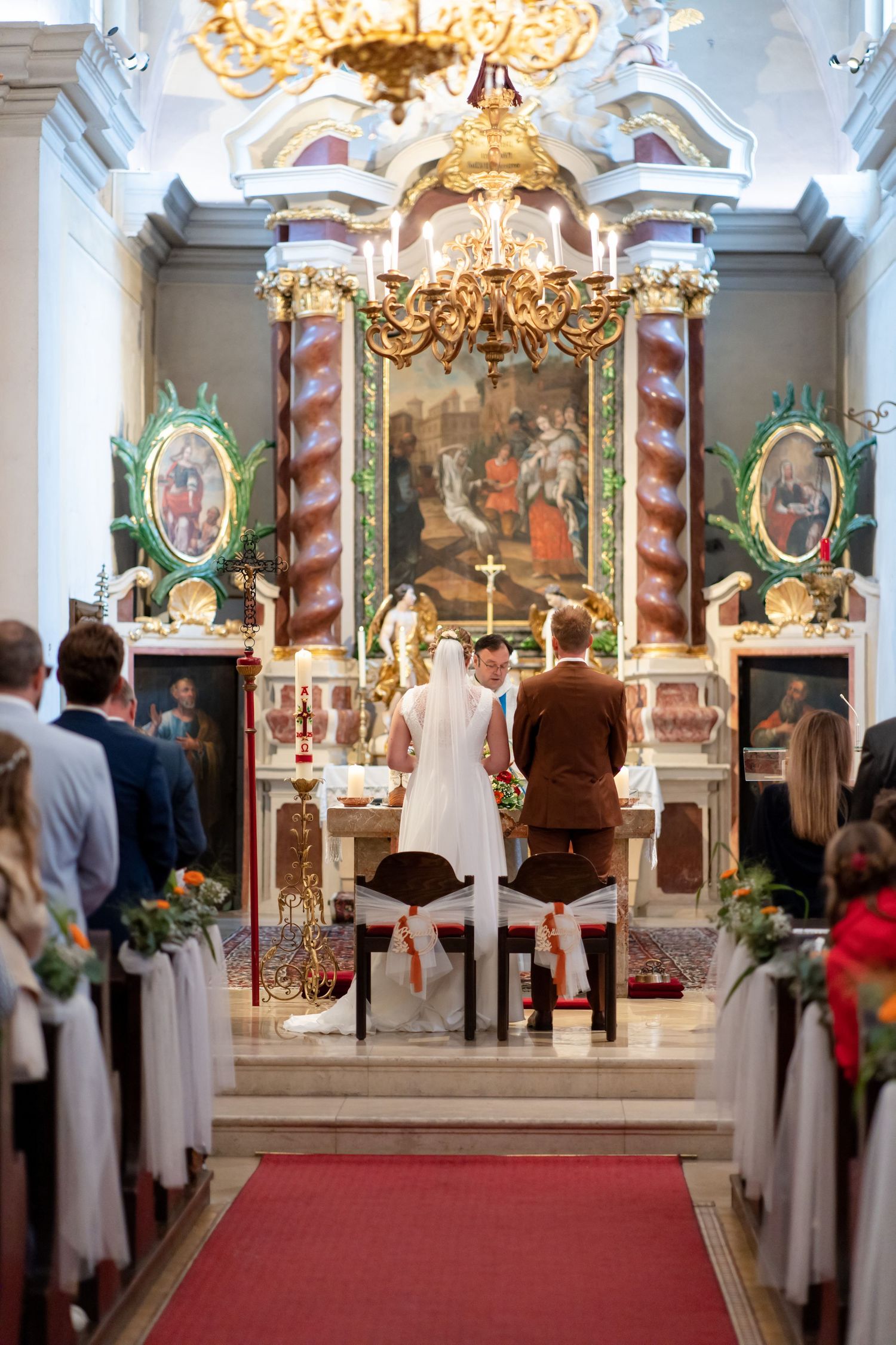 Interior of an ornate church with golden chandeliers and religious artwork during a wedding ceremony.