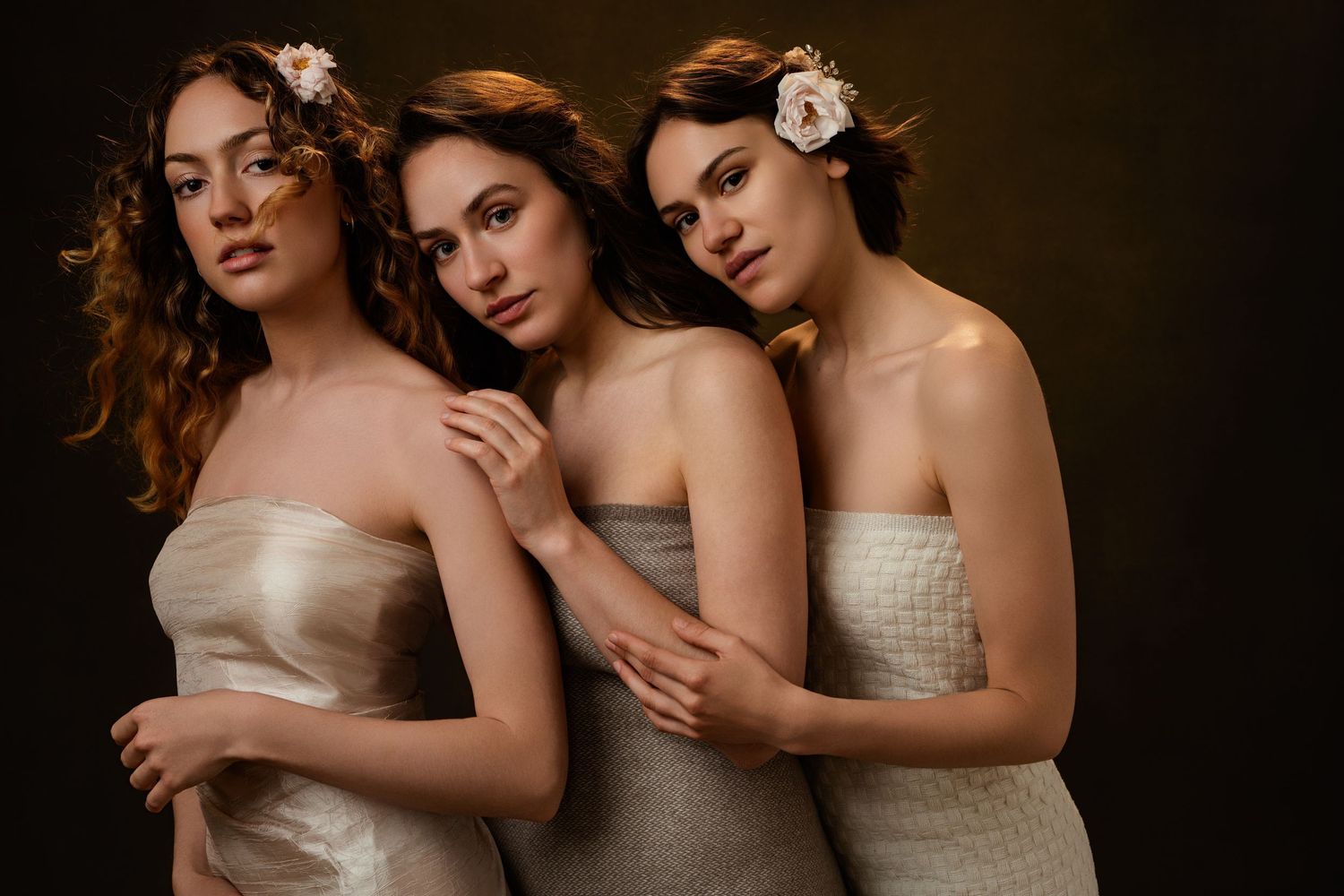 Three women in light colored dresses embrace intimately in a dramatic dark studio portrait.