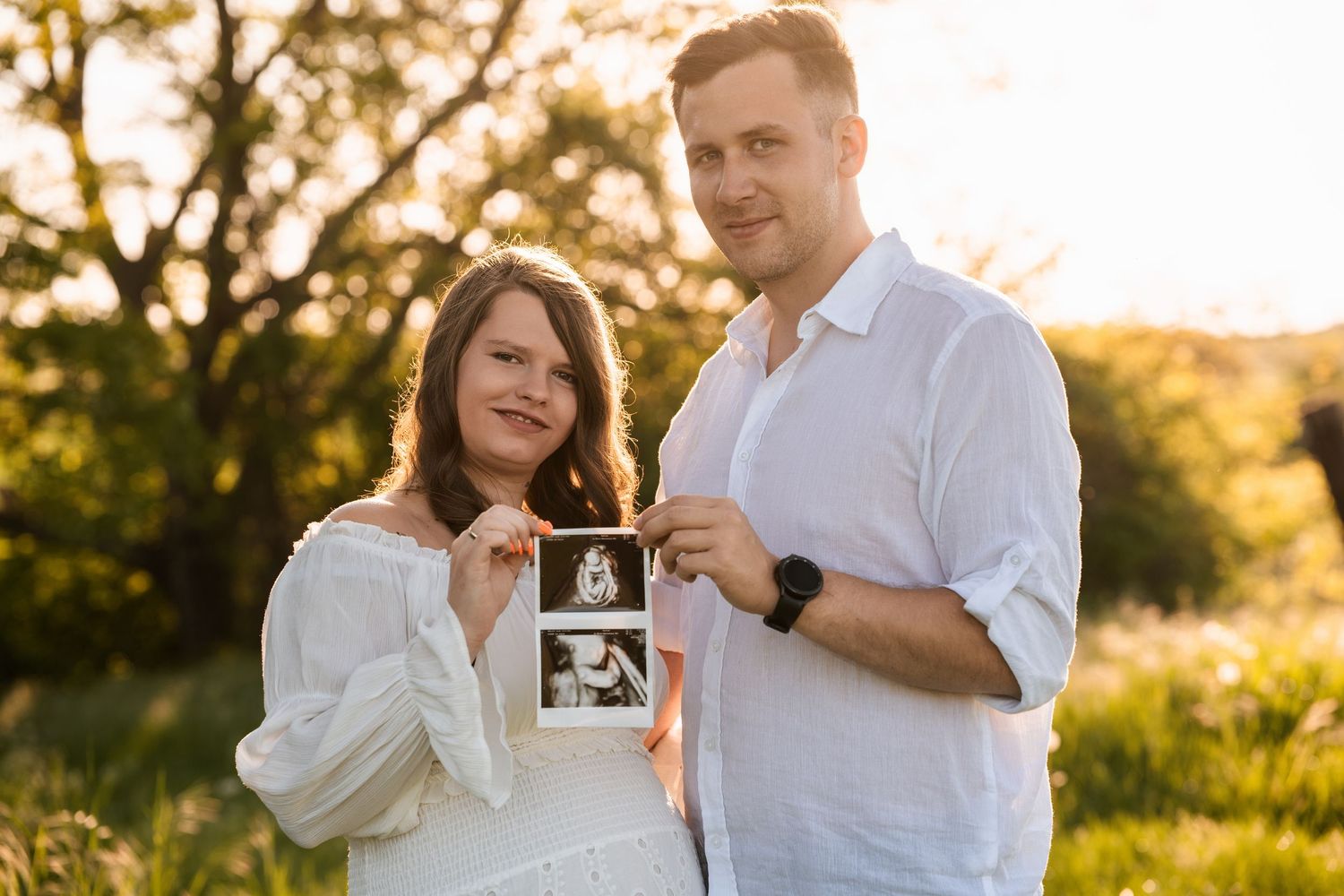 A couple in white outfits holds up ultrasound photos during a sunset maternity photoshoot in a natural outdoor setting.