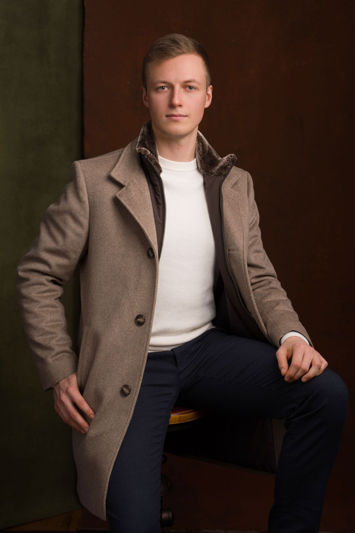 Young man in beige wool coat and white sweater seated confidently against dark studio background.