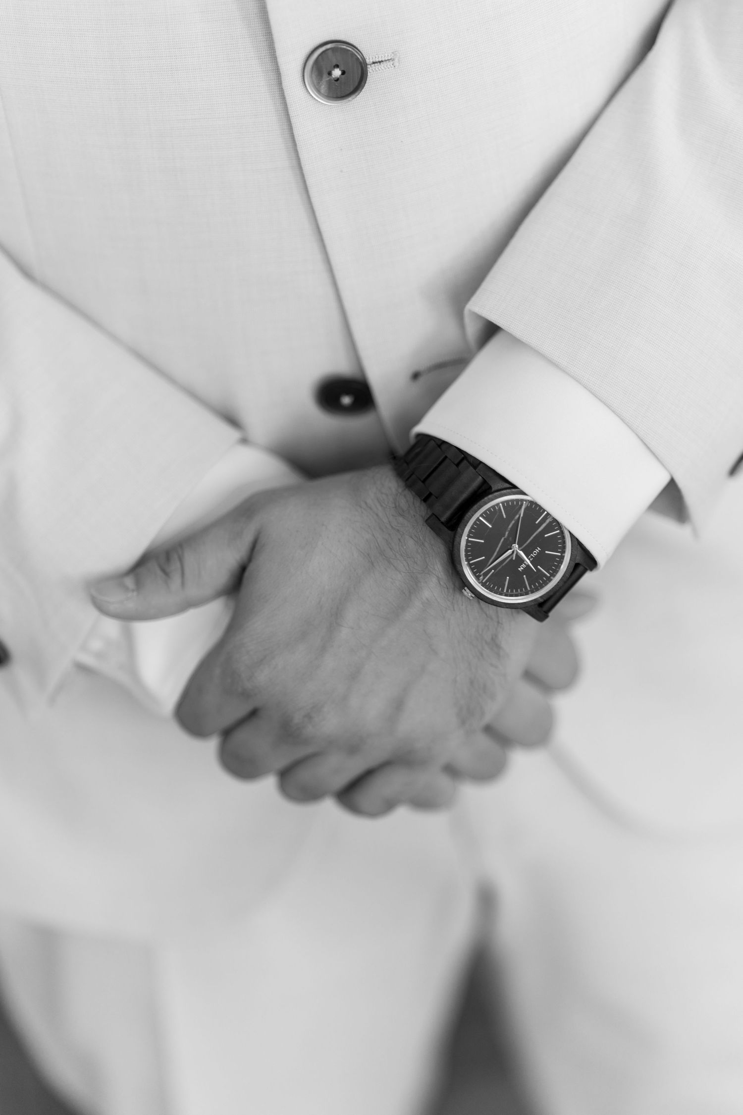 Groom in cream suit with black watch showing hands clasped in front.