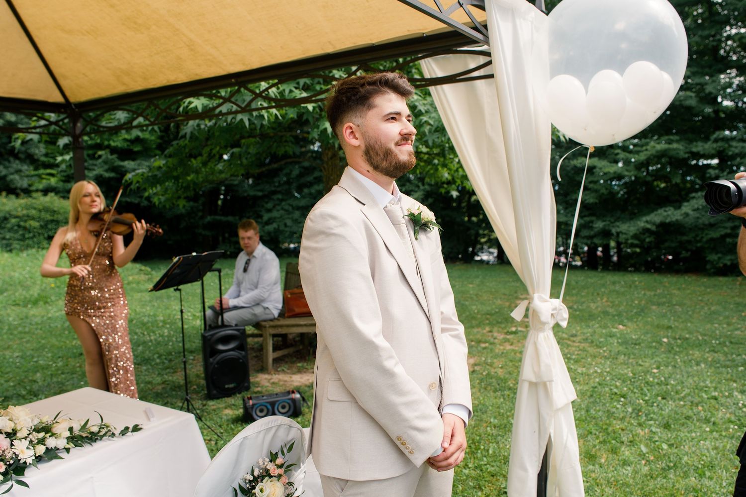 Groom in ivory suit stands at outdoor wedding altar with live musicians and white balloon decorations.