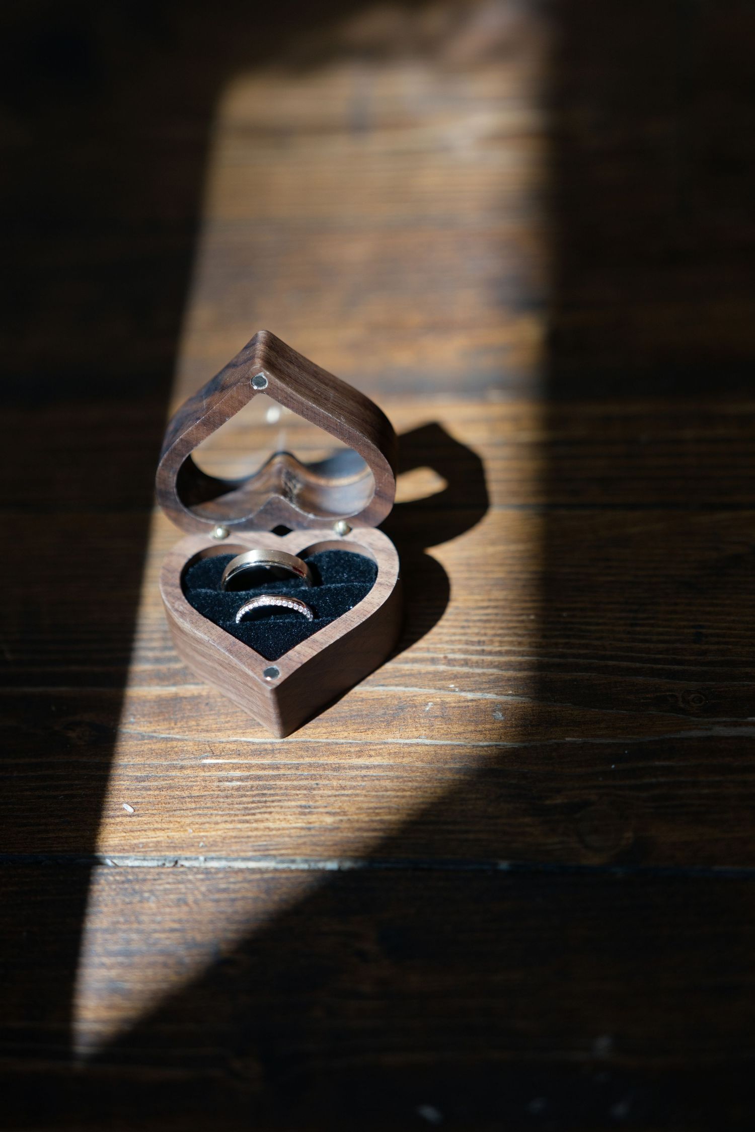 Heart-shaped wooden ring box with wedding bands illuminated by a dramatic beam of sunlight on a rustic wooden surface.