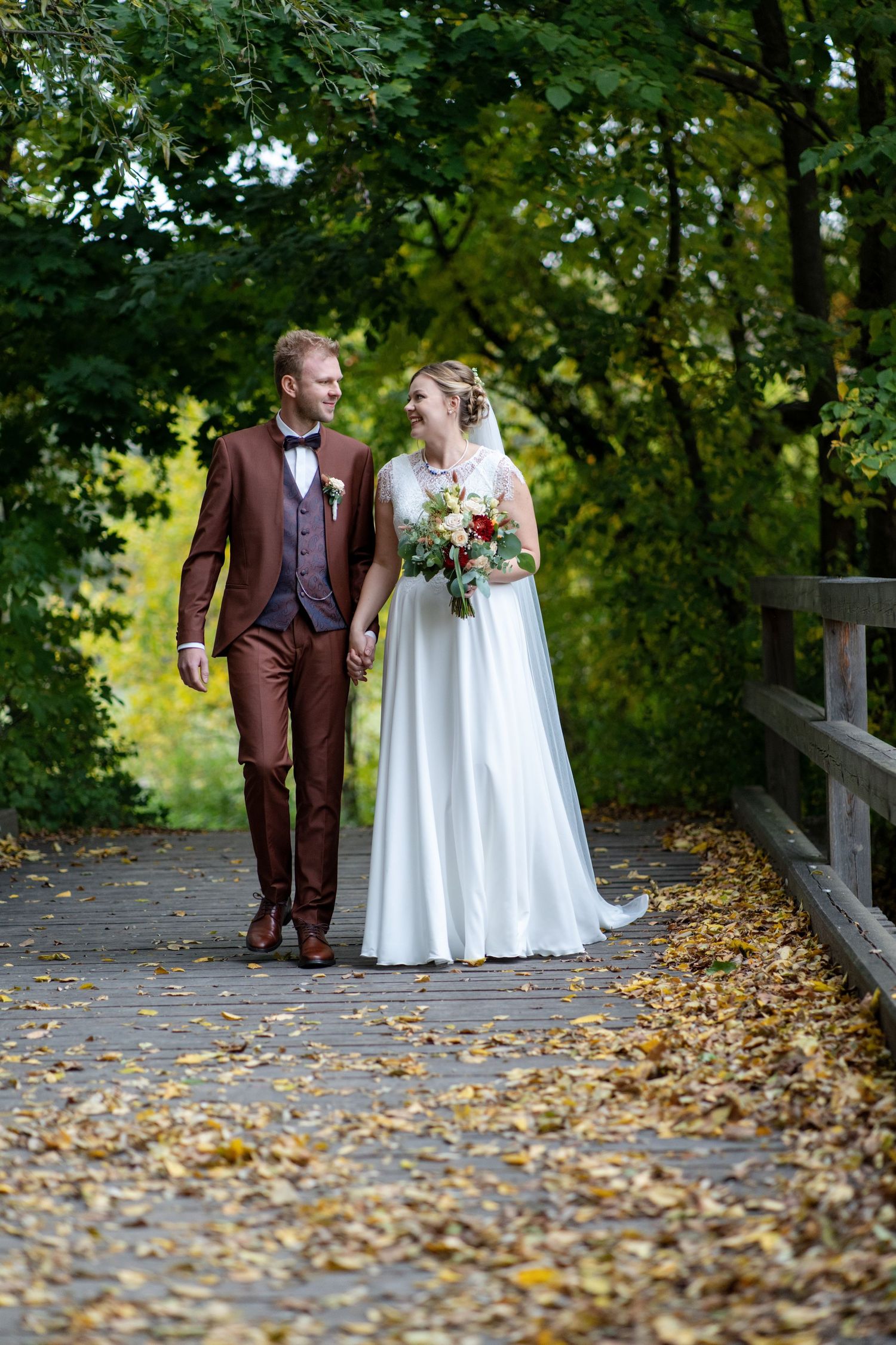 Newlyweds walking together on autumn path with fallen leaves and greenery.