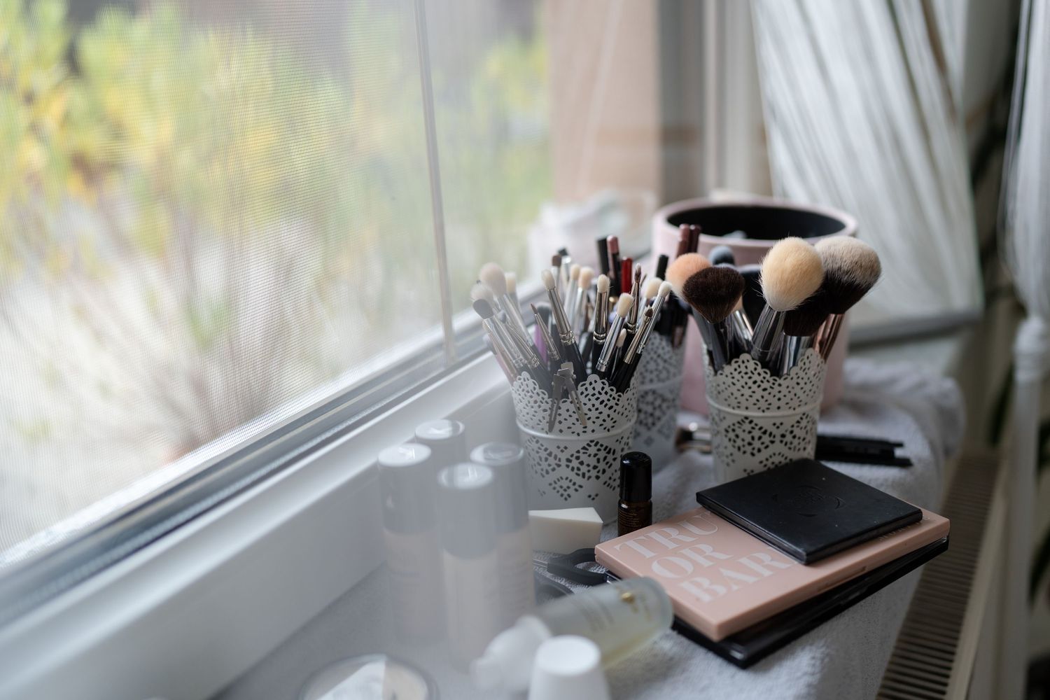 Organized desk setup near window with makeup brushes, plants, and white decor items arranged neatly.