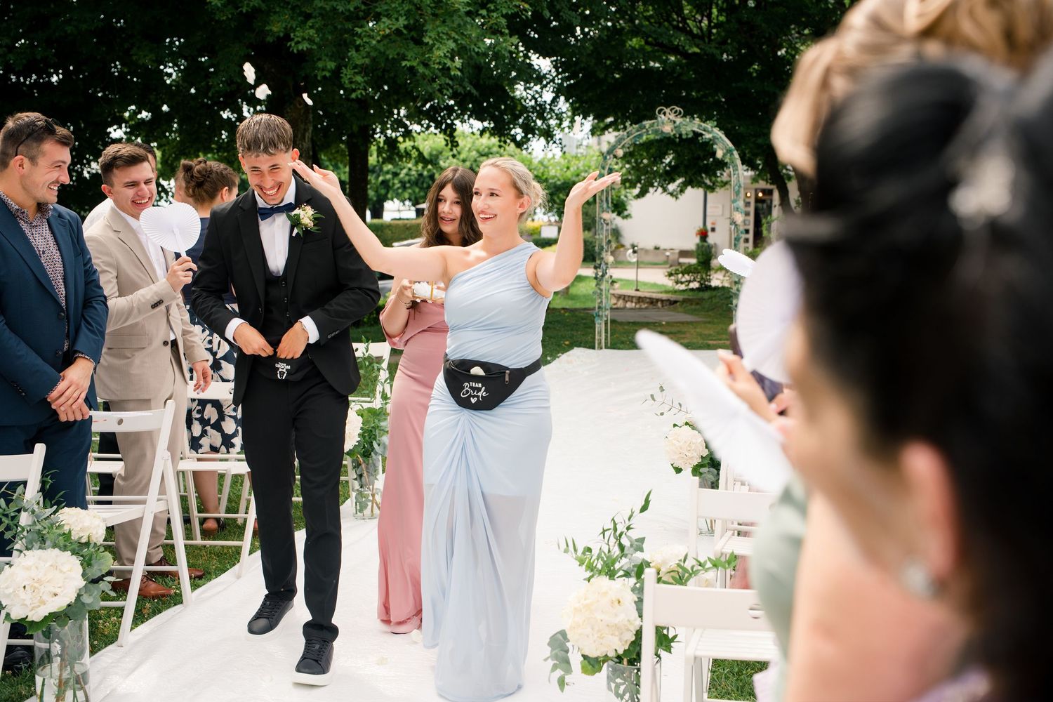 Wedding guests celebrate outdoors as a bridesmaid in blue dances joyfully with a groom in a black tuxedo.
