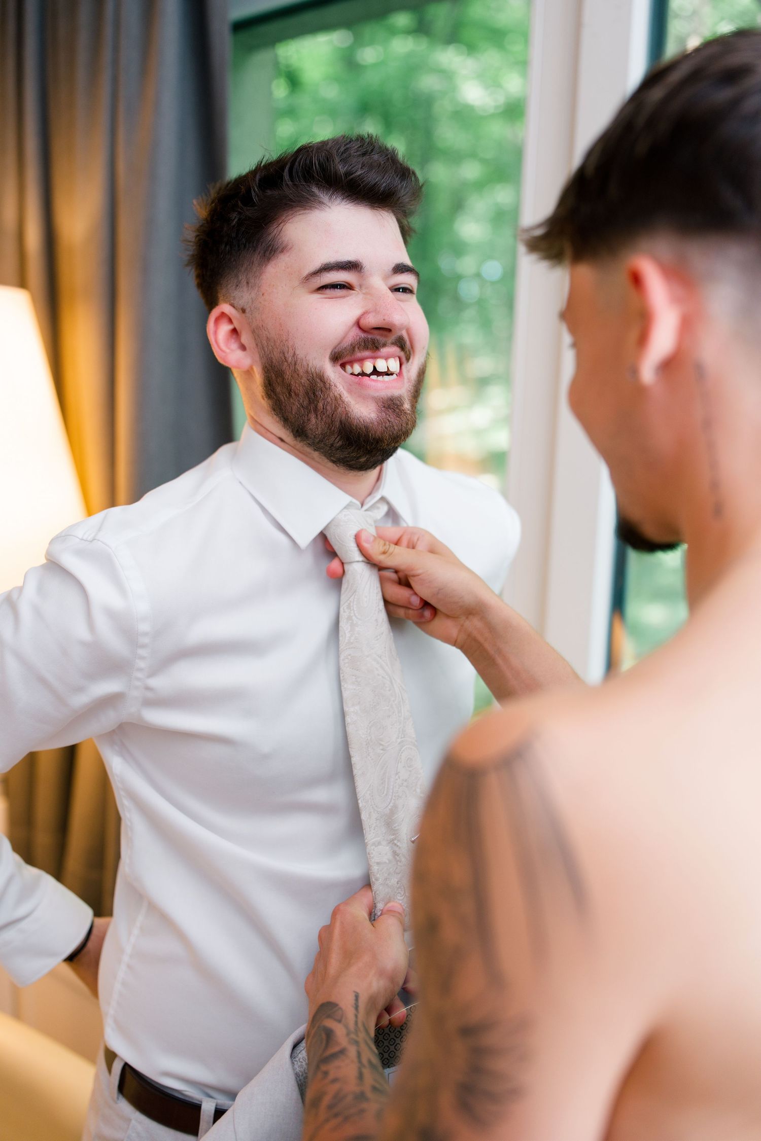 Smiling groom in white shirt gets tie adjusted by tattooed groomsman during wedding preparations.