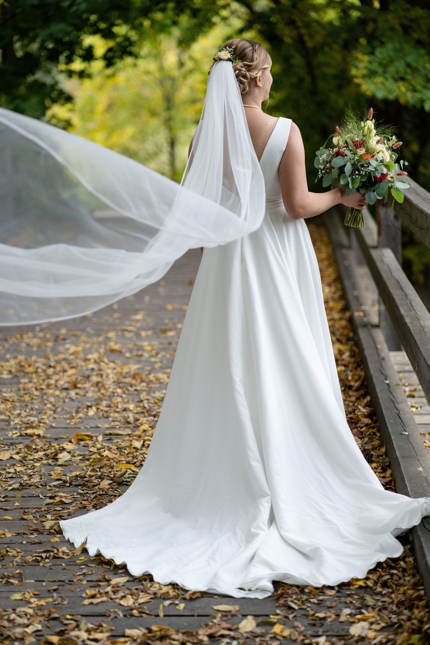 A flowing white wedding dress and veil captured from behind on an autumn path with falling leaves.
