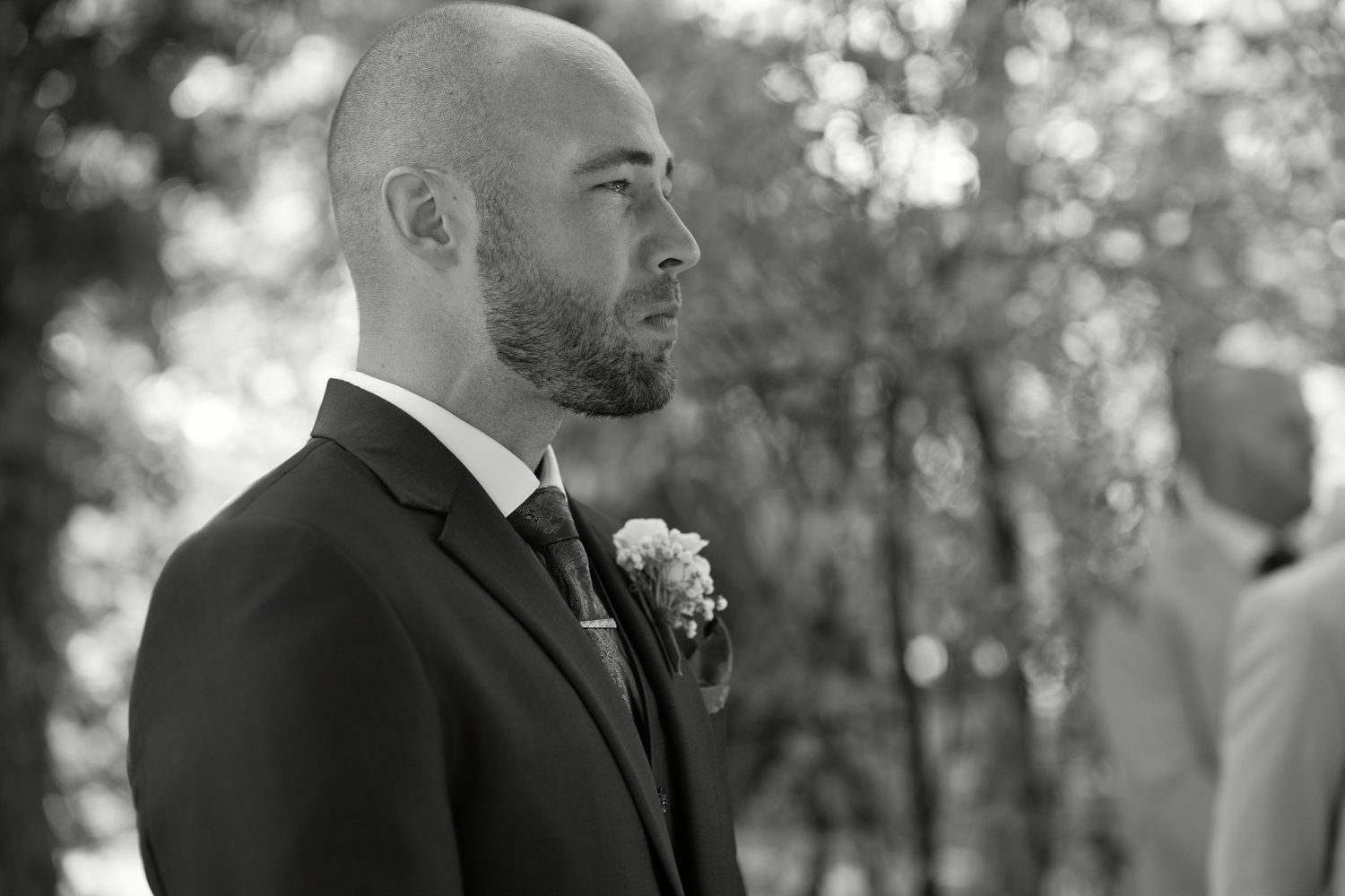 Black and white photo of a bald groom in a suit with boutonniere, looking emotional at an outdoor wedding ceremony.