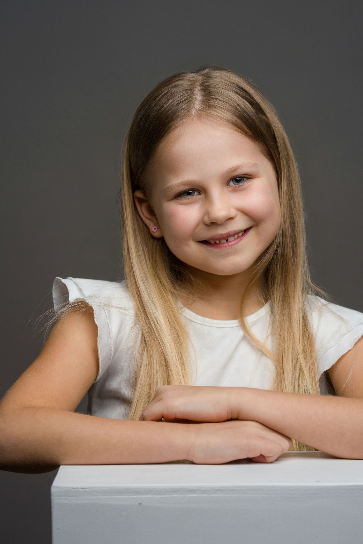 Child in white shirt leaning on table with a bright smile against dark backdrop.