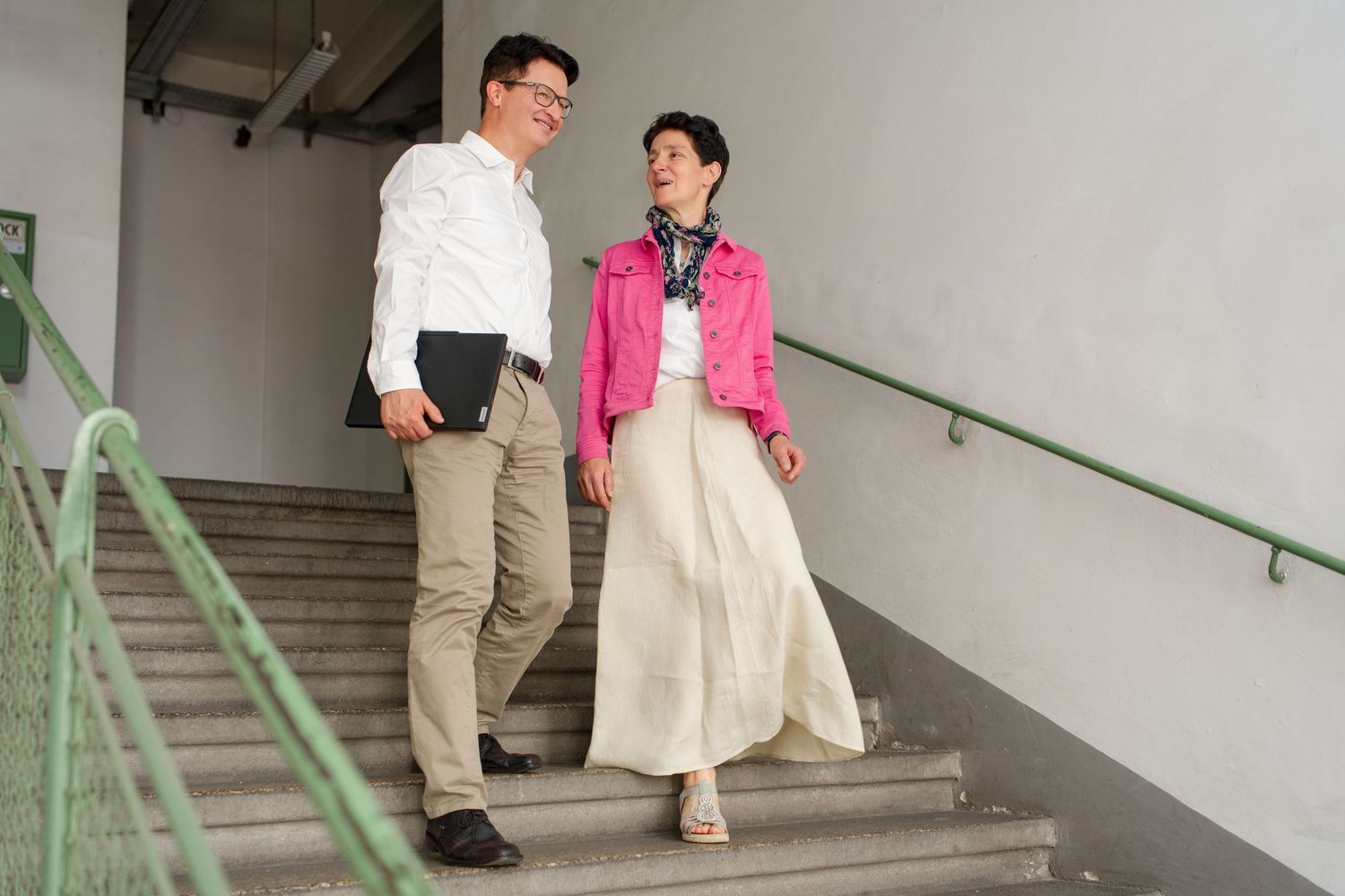 Man and woman walking down outdoor stairs, man in white shirt, woman in pink jacket and cream skirt.