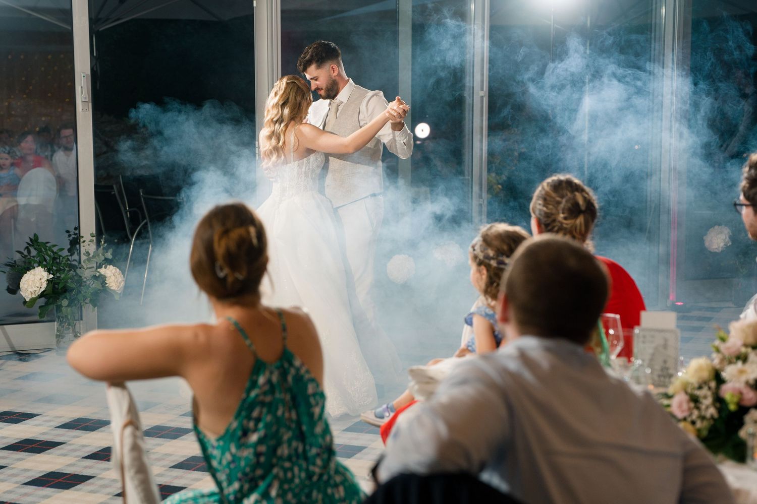 Bride and groom share first dance surrounded by smoke effects while wedding guests watch at elegant reception.