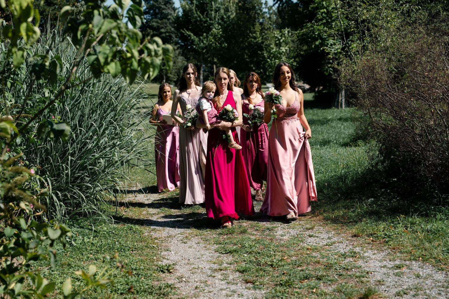 Bridesmaids in pink dresses walk along outdoor garden path.