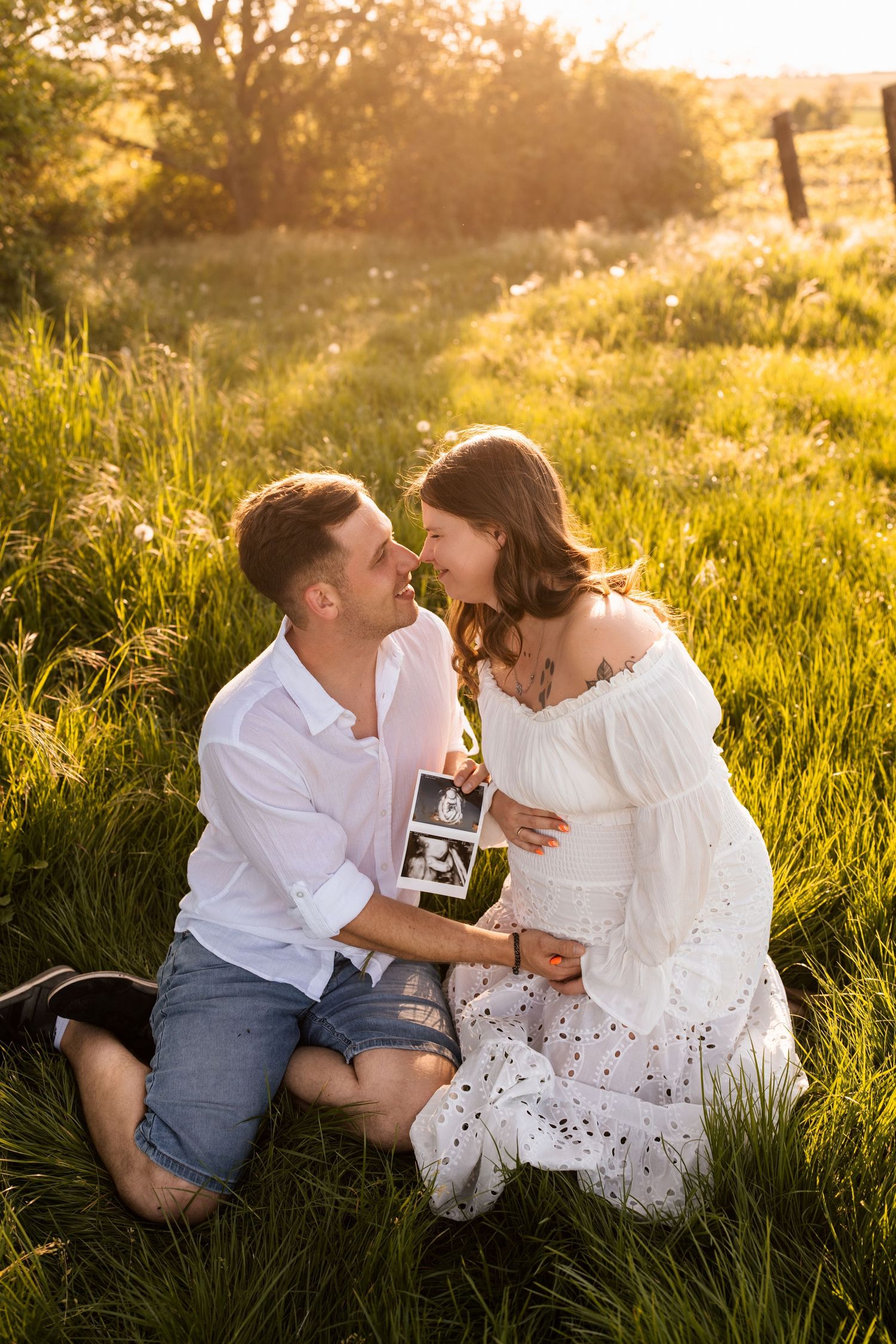 Couple sits in golden sunlit meadow sharing tender moments during sunset photoshoot.