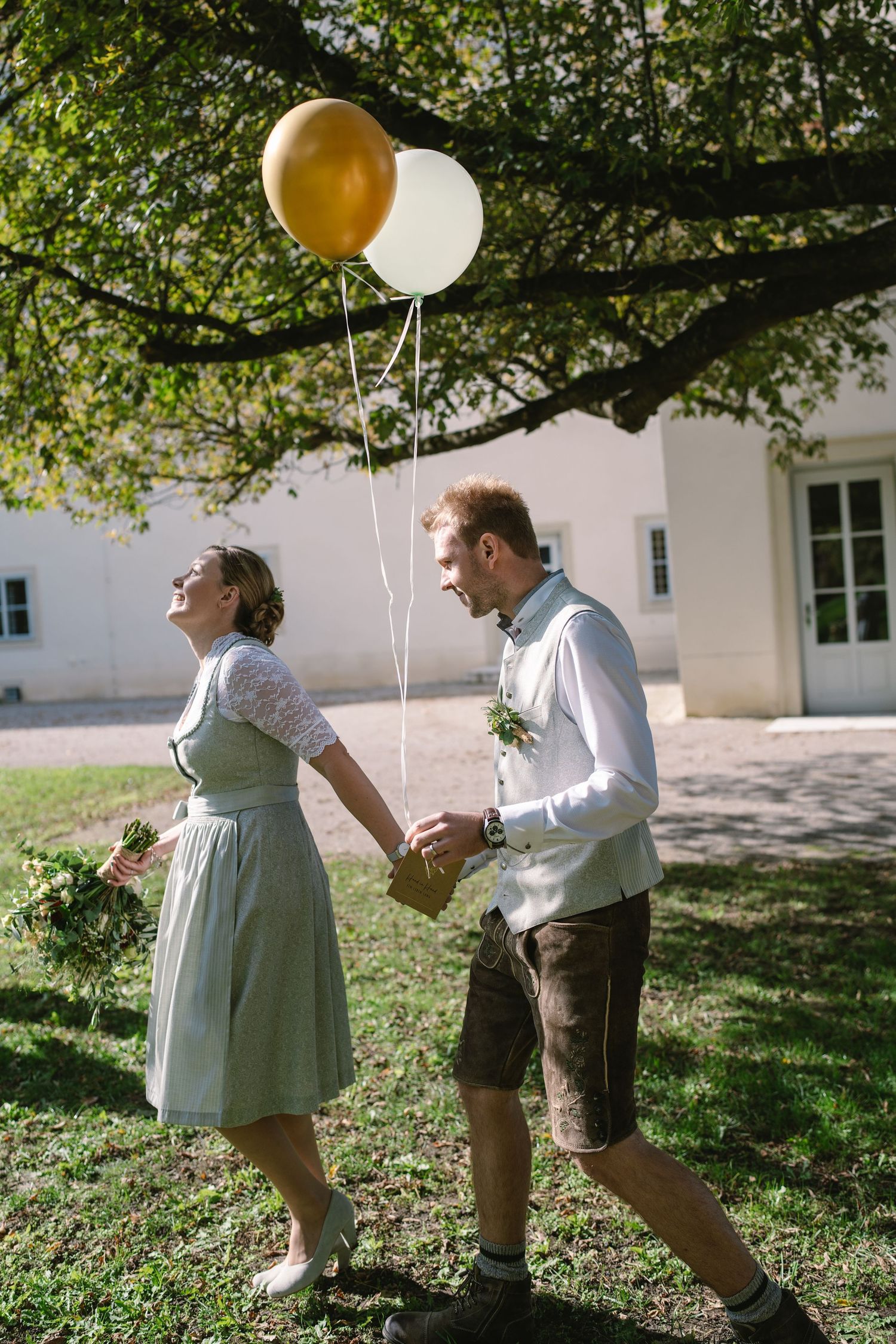 A couple in traditional Bavarian dress holds gold and white balloons while dancing outdoors on a sunny day.