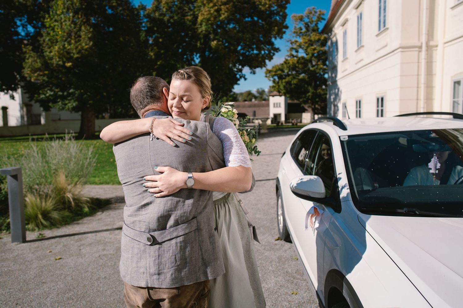 People embracing and hugging each other outdoors near parked cars on a sunny day.