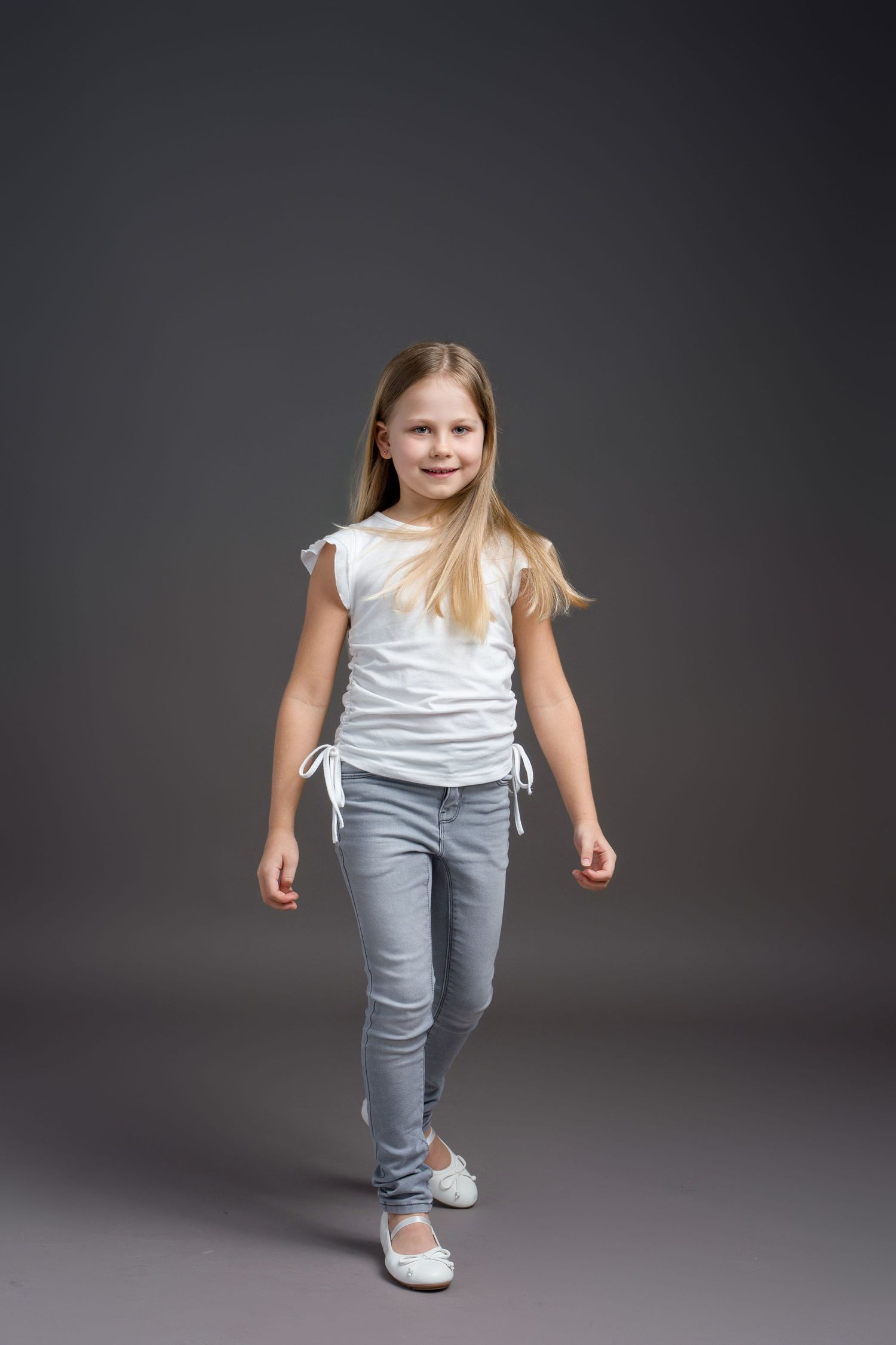 Child in casual white top and jeans walks confidently against dark studio backdrop.