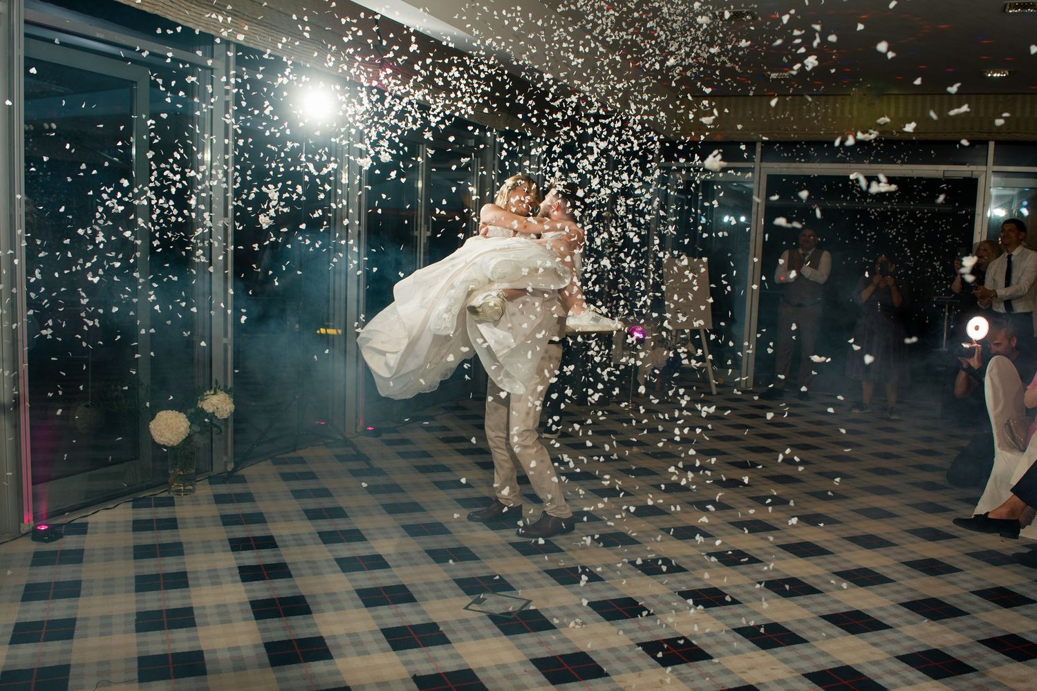 Groom lifting bride in wedding dress amid confetti shower on checkered floor at reception venue.