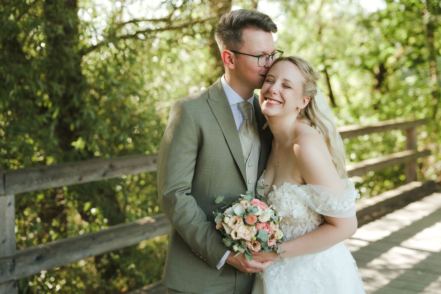Bride and groom embrace on a wooden bridge surrounded by lush greenery, bride holding a pink bouquet.