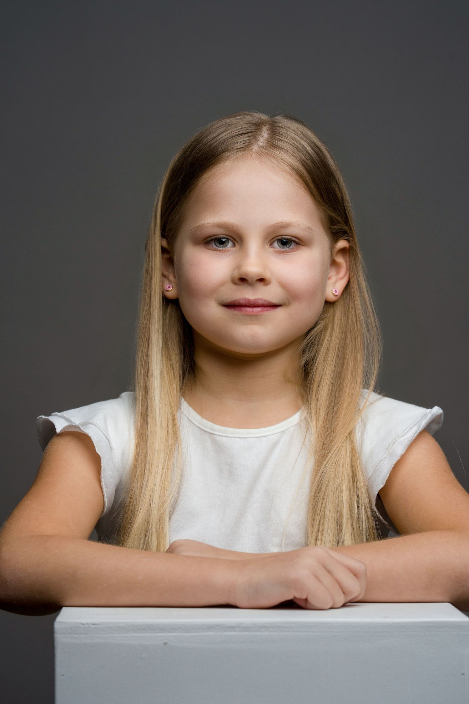 Young child with long blonde hair wearing a white shirt poses for a portrait against dark background.