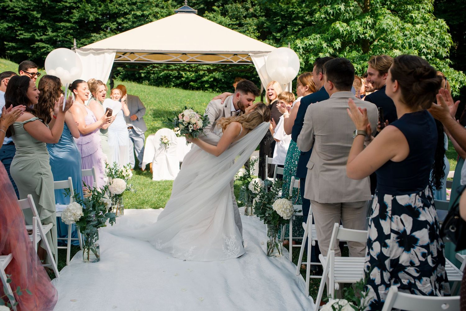 Newlywed couple sharing a kiss at outdoor wedding ceremony while guests applaud along white aisle.