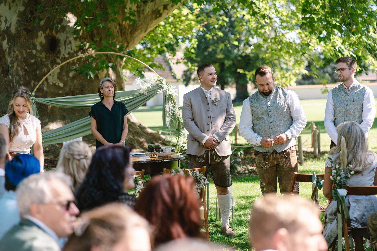 Wedding guests gathered outdoors under trees for garden ceremony.
