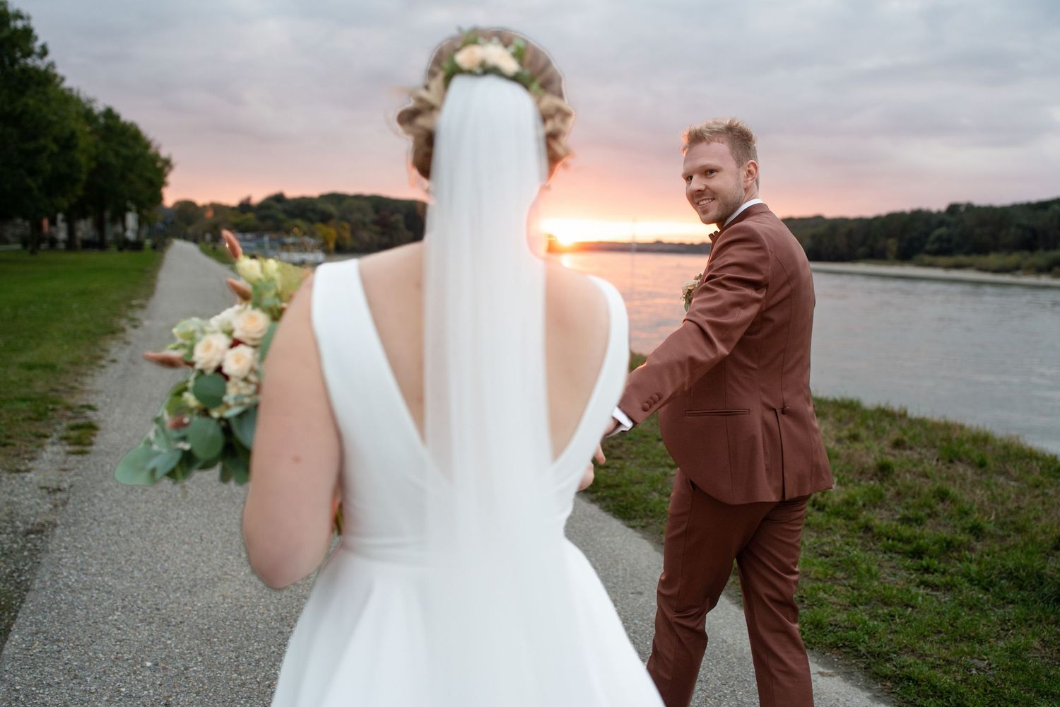 Wedding couple in brown suit and white dress pose at sunset near water.