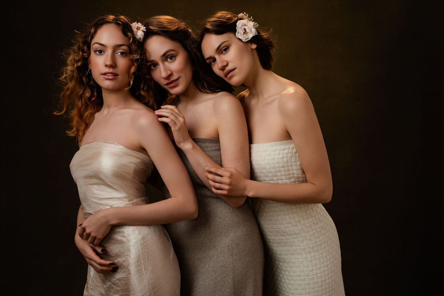 Three people in elegant cream-colored formal dresses pose together against a dark background.