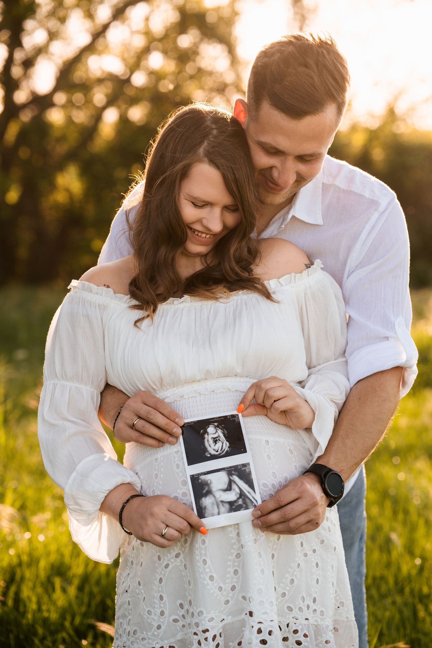 Couple embraces in golden sunlight while holding ultrasound photos in a grassy meadow during sunset.