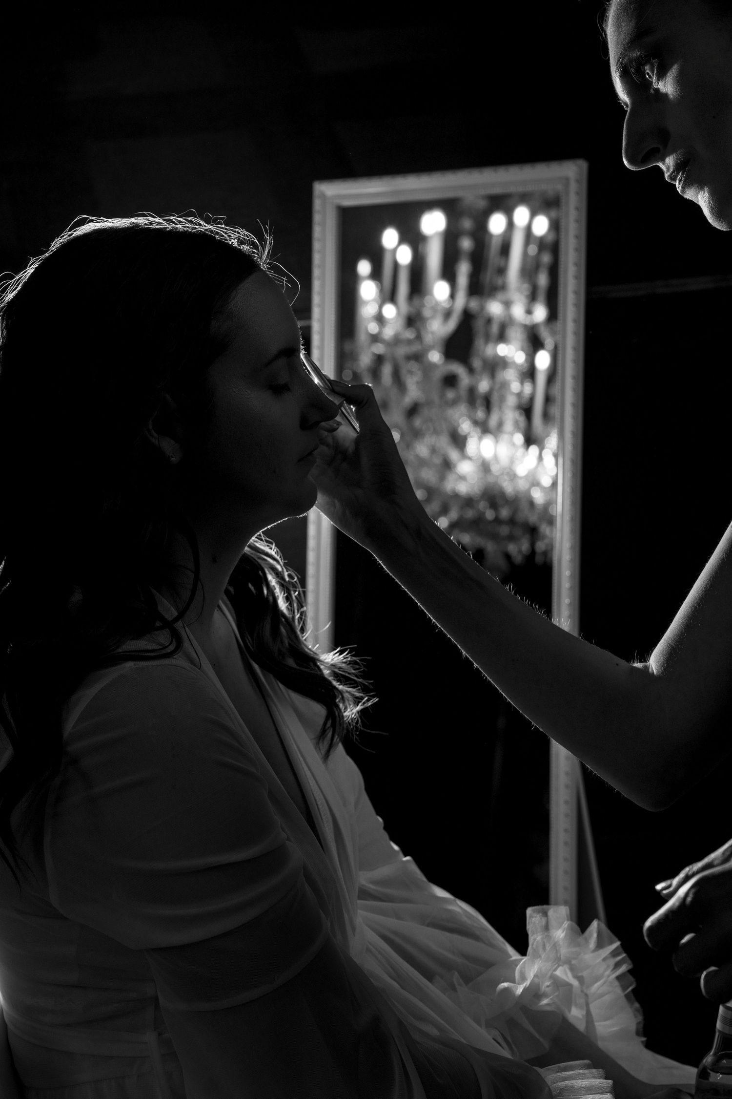 Black and white photo showing makeup application in a dimly lit room with chandelier reflection.