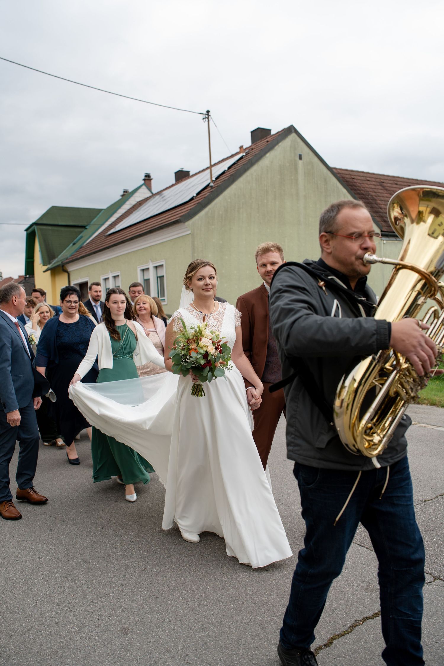 A wedding party walks down a street while musicians play brass instruments as the bride's dress flows in the wind.