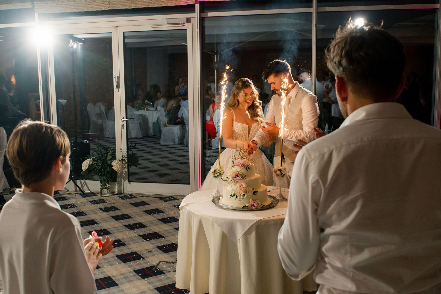 Bride and groom cutting wedding cake at elegant evening reception while guests watch.
