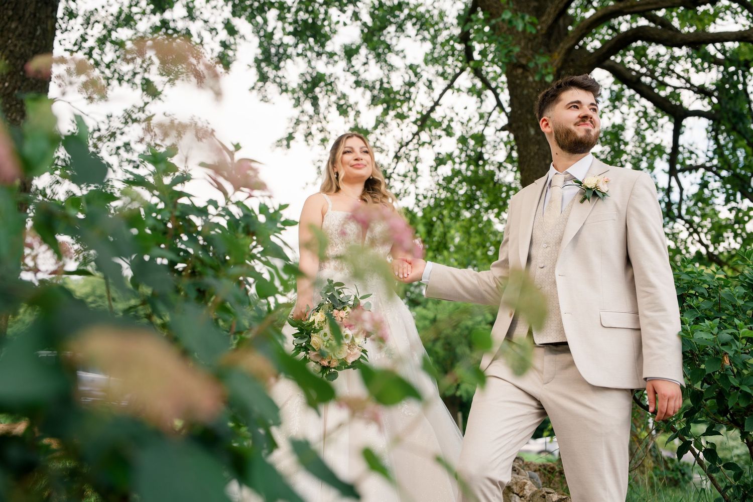 Bride and groom walking outdoors among lush green trees, groom in beige suit with boutonniere, bride in white gown holding flowers.